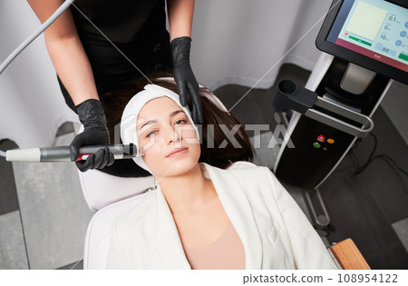 Top view of woman lying near cosmetology equipment and receiving skincare treatment. Female doctor beautician using radiofrequency microneedling device while performing face lifting procedure. 108954122