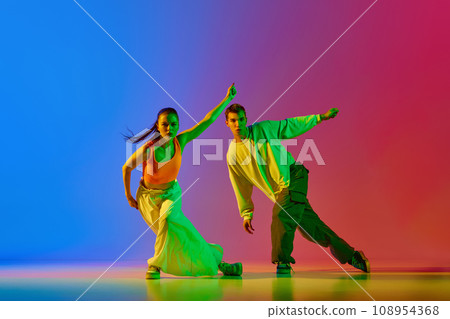 Coordinated movements. Young man and woman, street dancers in motion, dancing hip hop against blue red background in neon light 108954368