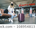 Young teenage girl is sitting at the airport waiting for her flight next to her suitcase. Girl wears a mask 108954813