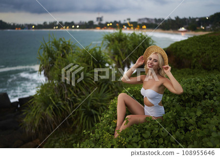 Blonde woman enjoys tropical beach vacation, wearing stylish white bikini and sun hat, posing amidst lush greenery with ocean waves and coastal city lights in background. Wellness travel inspiration. Blonde woman enjoys tropical beach vacation, wearing stylish white bikini and sun hat, posing amidst lush greenery with ocean waves and coastal city lights in background. Wellness travel inspiration. 108955646
