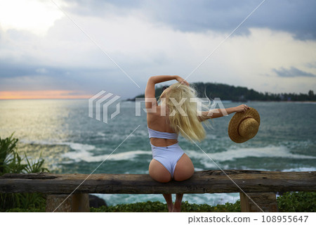 Woman in bikini with a beautiful physique sitting on beach looking at ocean view on sunset in swimwear. Slim figure girl enjoys sea vacation with straw hat in hands. Pretty slender fit blonde female. Woman in bikini with a beautiful physique sitting on beach looking at ocean view on sunset in swimwear. Slim figure girl enjoys sea vacation with straw hat in hands. Pretty slender fit blonde female. 108955647