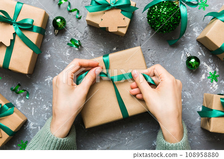 Woman is packing surprise gift for Christmas holidays, sitting at workplace with craft tools, top view. Woman wrapping Christmas gift at colored table 108958880