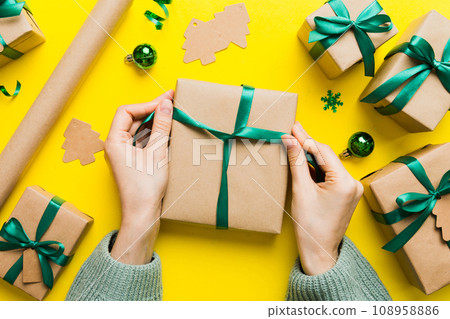 Woman is packing surprise gift for Christmas holidays, sitting at workplace with craft tools, top view. Woman wrapping Christmas gift at colored table 108958886