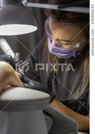 Manicurist at work. Professional manicurist removes old worn nail design from nails of client using modern electric drill. Preparing fingernails for making fresh manicure and applying new nail design 108960244