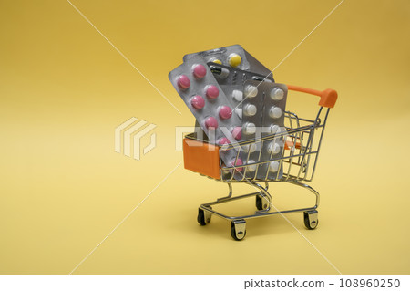 Many medicine pills, tablets in a small supermarket shopping cart on yellow background. concept shopping medicine. Pharmacy theme, capsular tablets with medicinal vitamins Many medicine pills, tablets in a small supermarket shopping cart on yellow background. concept shopping medicine. Pharmacy theme, capsular tablets with medicinal vitamins 108960250