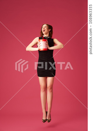 Happy young girl in elegant black dress holding present, celebrating against pink studio background. Birthday Happy young girl in elegant black dress holding present, celebrating against pink studio background. Birthday 108963355