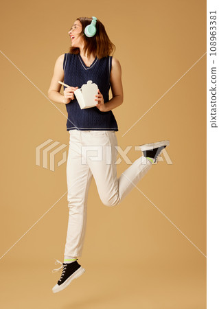 Positive young girl in headphones eating noodles with chopsticks against beige studio background. Dinner to go. Positive young girl in headphones eating noodles with chopsticks against beige studio background. Dinner to go. 108963381