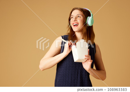 Positive young girl in headphones eating noodles with chopsticks against beige studio background. Dinner to go. 108963383