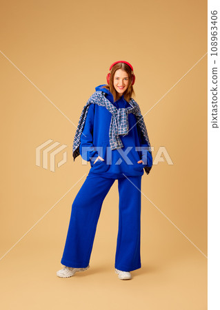 Full-length image of young girl in stylish blue clothes and shirt, listening to music in headphones against beige studio background 108963406