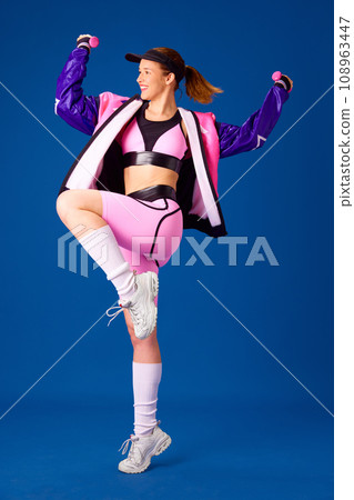 Positive, smiling young girl in colorful sportswear standing with dumbbells against blue studio background Positive, smiling young girl in colorful sportswear standing with dumbbells against blue studio background 108963447