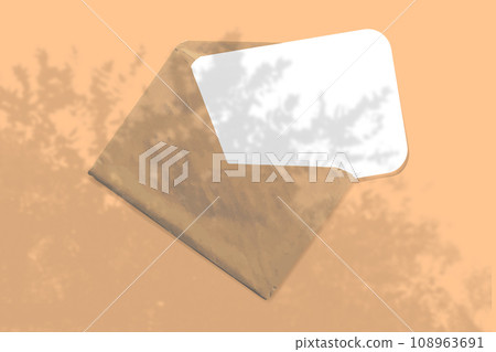 An envelope with sheet of textured white paper on yellow table background. Mockup with an overlay of tropical plant shadows. Natural light casts shadows from the leaves of a tree branch. Horizontal 108963691