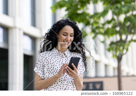 A young beautiful woman walks through the city with a phone in her hands, smiles contentedly, uses a smartphone application, browses Internet pages, types a text message, social networks. 108965122