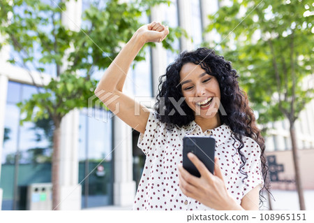 Young joyful woman winner received online notification on phone, Hispanic woman with curly hair celebrating success and triumph walking in city near office building outside. Young joyful woman winner received online notification on phone, Hispanic woman with curly hair celebrating success and triumph walking in city near office building outside. 108965151