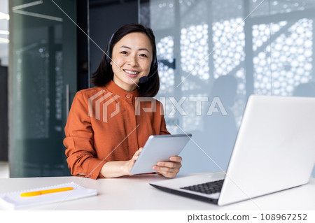 Portrait of successful satisfied female online customer service support worker, Asian business woman looking at camera, holding tablet computer, using headset and laptop for video call. 108967252