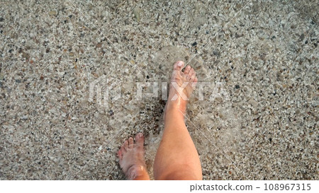 Male feet walking over sandy ocean beach and pebbles through calm clear sea water 108967315