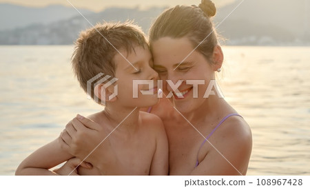 Portrait of loving mother and son hugging each other while relaxing on the sea against sunset light rays Portrait of loving mother and son hugging each other while relaxing on the sea against sunset light rays 108967428