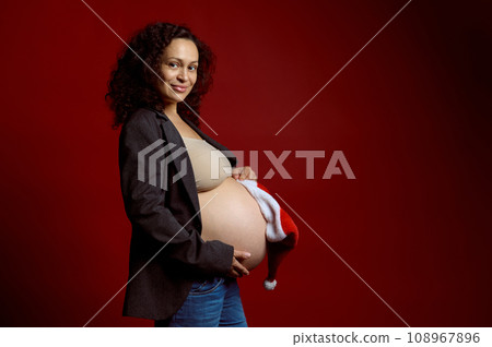 Confident gravid female holding Santa hat over her naked big belly in the third trimester of pregnancy, smiles at camera Confident gravid female holding Santa hat over her naked big belly in the third trimester of pregnancy, smiles at camera 108967896