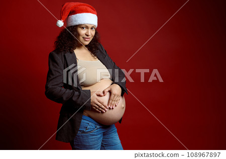 Charming pregnant woman in Santa hat, forming a shape of heart over her naked belly, smiling at camera, red background Charming pregnant woman in Santa hat, forming a shape of heart over her naked belly, smiling at camera, red background 108967897