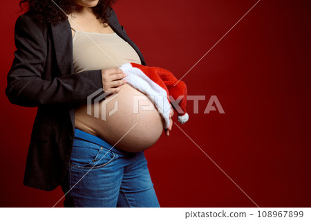 Closeup belly of gravid woman, expectant mom holding Santa hat on her pregnant belly, isolated on red studio background 108967899