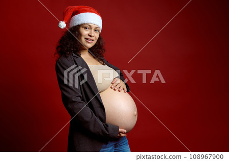 Charming pregnant woman in red Santa hat, gently stroking her big belly, smiling looking at camera isolated red backdrop Charming pregnant woman in red Santa hat, gently stroking her big belly, smiling looking at camera isolated red backdrop 108967900