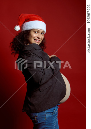 Authentic pregnant woman in Santa hat, smiles at camera, expressing positive emotions, isolated red studio background 108967901