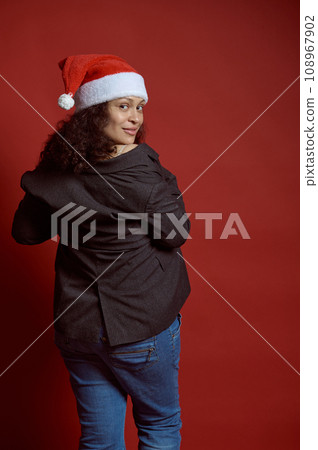 Back view beautiful ethnic young adult pregnant woman wearing Santa hat, smiles cutely looking at camera, red background Back view beautiful ethnic young adult pregnant woman wearing Santa hat, smiles cutely looking at camera, red background 108967902