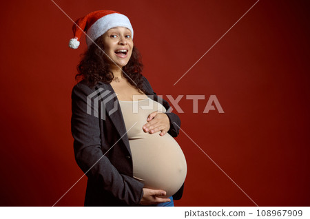 Pregnant woman wearing Santa hat, holds belly, smiles looking at camera, isolated red studio background. Merry Christmas Pregnant woman wearing Santa hat, holds belly, smiles looking at camera, isolated red studio background. Merry Christmas 108967909