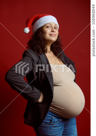 Studio portrait of emotional charismatic pregnant woman in Santa hat, smiling looking at camera, isolated red background Studio portrait of emotional charismatic pregnant woman in Santa hat, smiling looking at camera, isolated red background 108968049