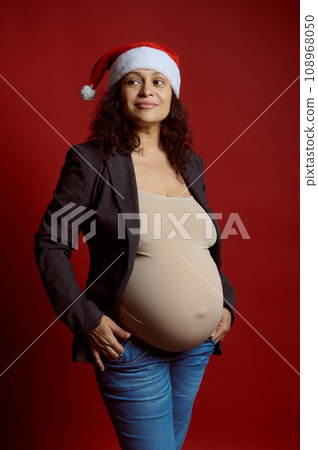 Studio portrait of emotional charismatic pregnant woman in Santa hat, smiling looking aside, isolated on red background 108968050
