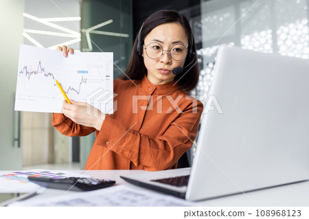 Serious concentrated thinking woman on online presentation, video meeting, business woman focused showing to camera laptop income graph of new project, working with headset phone inside office. Serious concentrated thinking woman on online presentation, video meeting, business woman focused showing to camera laptop income graph of new project, working with headset phone inside office. 108968123