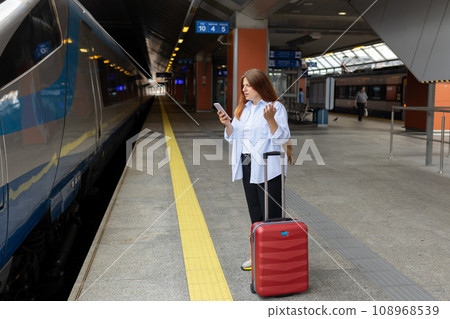 Young refugee redhhead woman crying and waiting train on station platform, she lost and using smart phone. Railroad transport concept, full body 108968539