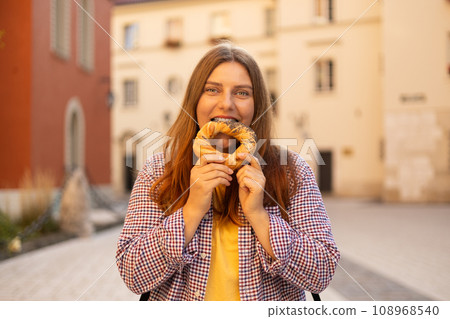 Attractive young female 30s tourist with backpack is holding prezel, traditional polish snack on the Market square in Krakow. Traveling Europe in spring. Selective focus. High quality photo 108968540