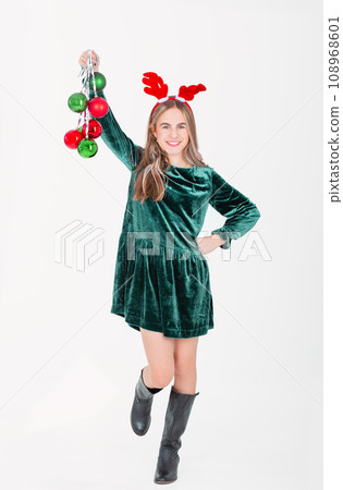 Festively dressed cute girl wearing reindeer antlers holding Christmas decorations on isolated white background Festively dressed cute girl wearing reindeer antlers holding Christmas decorations on isolated white background 108968601