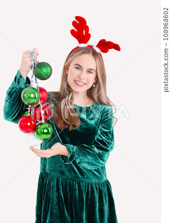 Festively dressed cute girl wearing reindeer antlers holding Christmas decorations on isolated white background Festively dressed cute girl wearing reindeer antlers holding Christmas decorations on isolated white background 108968602