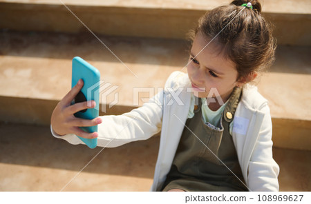 Adorable elementary age Caucasian child girl holding mobile phone and making selfie outdoors 108969627