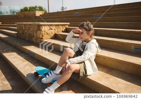 Cute little kid girl with skateboard sitting on steps outdoors and dreamily looking into the distance. Cute little kid girl with skateboard sitting on steps outdoors and dreamily looking into the distance. 108969642