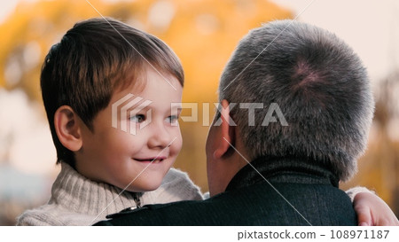 Father holds smiling son in arms with warmth while stands in park. Father amid ambiance of park holds son creating moment. Father in setting of park holding son in arms underscoring beauty of season 108971187