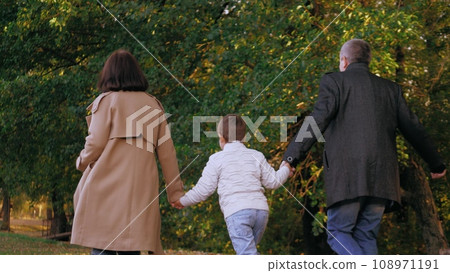 Happy family runs across autumn park joining hands. Family of parents with little son runs along path past yellowed trees. Family of mother and father spends time with beloved son having fun in park Happy family runs across autumn park joining hands. Family of parents with little son runs along path past yellowed trees. Family of mother and father spends time with beloved son having fun in park 108971191