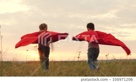 Children races on field in capes resembling superheroes. Children wearing capes dashes across field with energy at dusk. Children team playing superheroes runs on field while capes flutter in wind 108971206