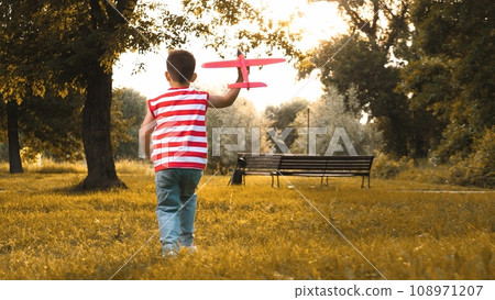 Schoolboy gleefully dashes across park with boundless energy. Schoolboy imitating pilot runs with excitement holding toy airplane. Schoolboy in T-shirt dashes through park holding toy airplane Schoolboy gleefully dashes across park with boundless energy. Schoolboy imitating pilot runs with excitement holding toy airplane. Schoolboy in T-shirt dashes through park holding toy airplane 108971207
