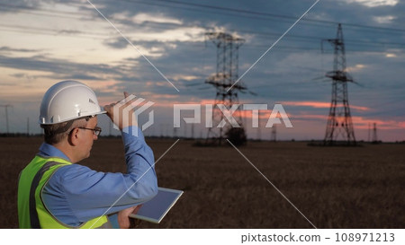Electrician holds tablet and adjusts helmet working at power distributions substation in sunset field. Engineer with tablet setups power generation facility at sunset. Electrical power manager 108971213