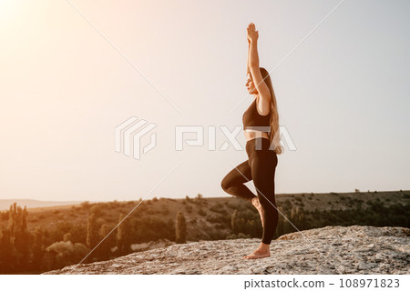 Fitness woman. Happy middle-aged fitness woman doing stretching and pilates on a rock near forest at sunset. Female fitness yoga routine. Healthy lifestyle with focus on well-being and relaxation. 108971823
