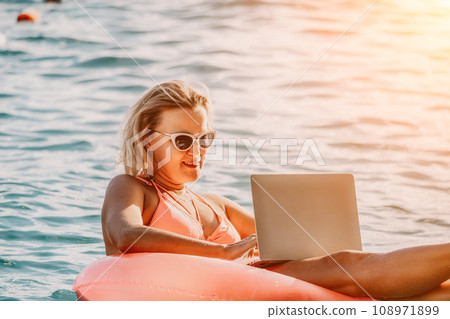 Woman works on laptop in sea. Freelancer, young blond woman in sunglases floating on an inflatable big pink donut with a laptop in the sea at sunset. Freelance, travel and holidays concept. 108971899