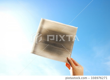 Car air conditioner filter clogged and dirty in a hand on blue sky background with sunlight Car air conditioner filter clogged and dirty in a hand on blue sky background with sunlight 108976271