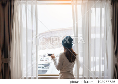 young woman in sweater with cup of coffee looking through the window in winter season, happy female enjoying snowfall outdoor view at apartment or home in the morning. Waking and Relaxing concept 108977786