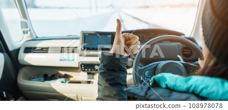 Happy Traveler driving car on snowy road and gesture finger up, woman Tourist enjoying snow forest view from the car in winter season. Winter travel, Road trip, Exploring and Vacation concepts 108978078