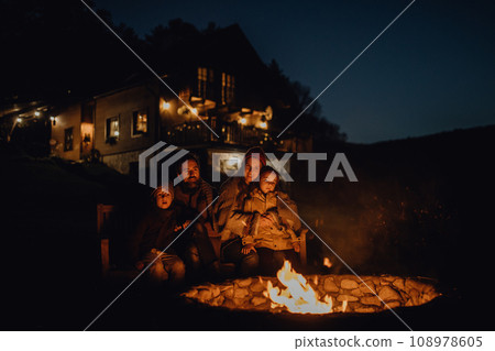 Family is warming up by an outdoor fire pit, sitting in the evening warmth of the fire, spending quality family time together. Family is warming up by an outdoor fire pit, sitting in the evening warmth of the fire, spending quality family time together. 108978605
