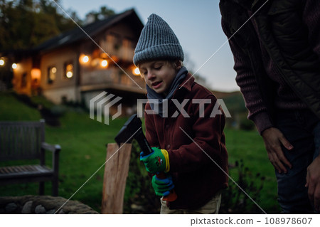 Young boy helping chop wood outdoors, preparing firewood for heating in indoor fireplace. Young boy helping chop wood outdoors, preparing firewood for heating in indoor fireplace. 108978607