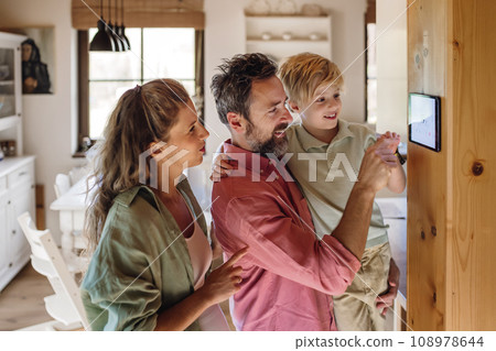 Family looking at smart thermostat, adjusting, lowering heating temperature at home. Concept of sustainable, efficient, and smart technology in home heating and thermostats. Family looking at smart thermostat, adjusting, lowering heating temperature at home. Concept of sustainable, efficient, and smart technology in home heating and thermostats. 108978644