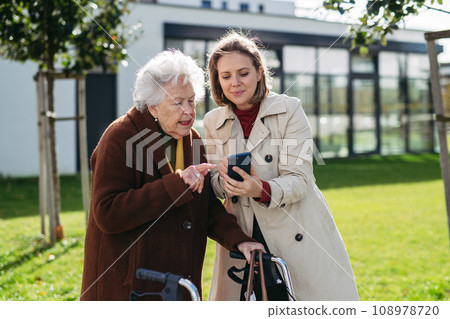 Mature granddaughter teaching grandmother how to use smartphone. Caregiver helping senior lady shopping online on smartphone. 108978720